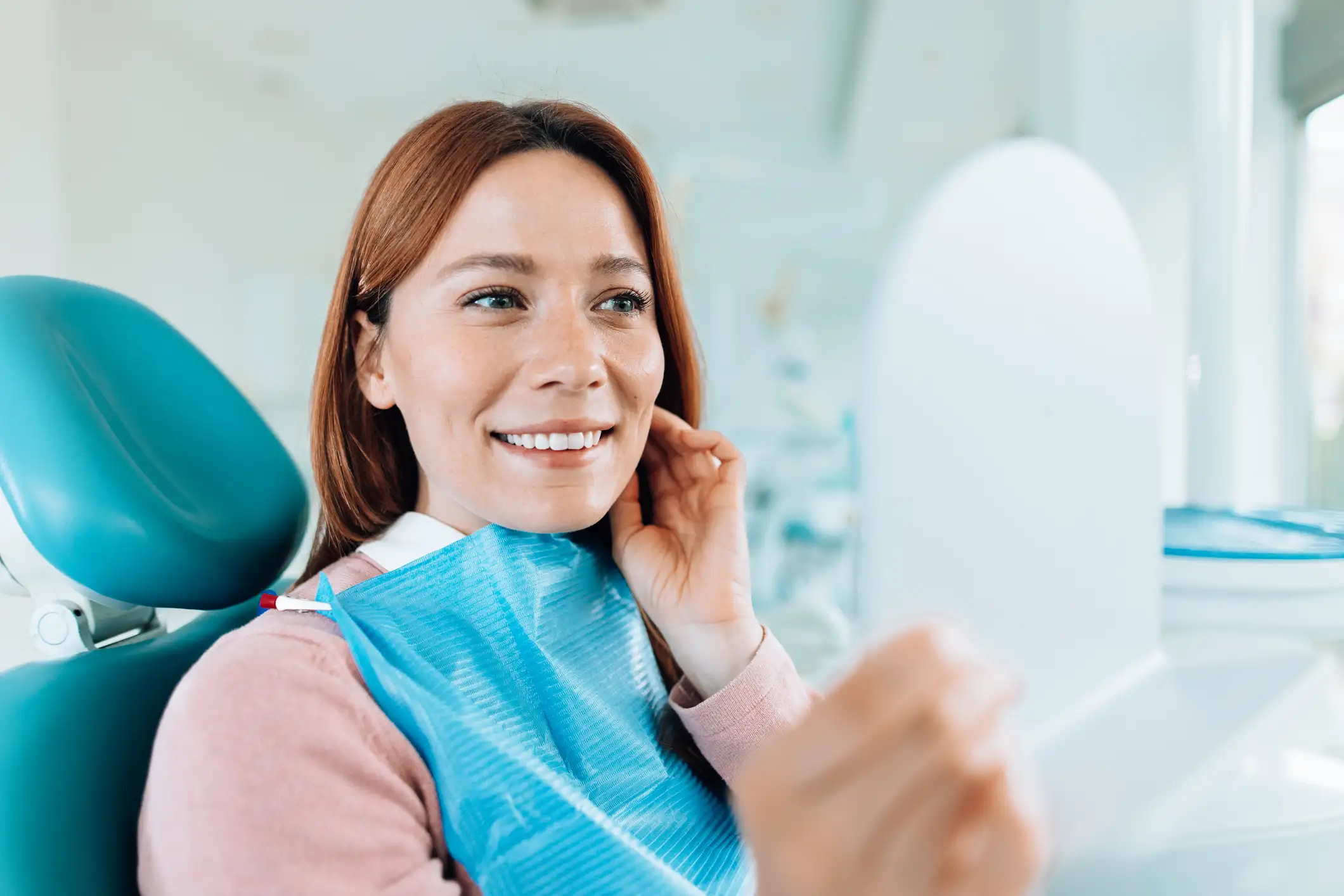 woman holding a mirror in a dentist's chair looking at her new, perfect smile